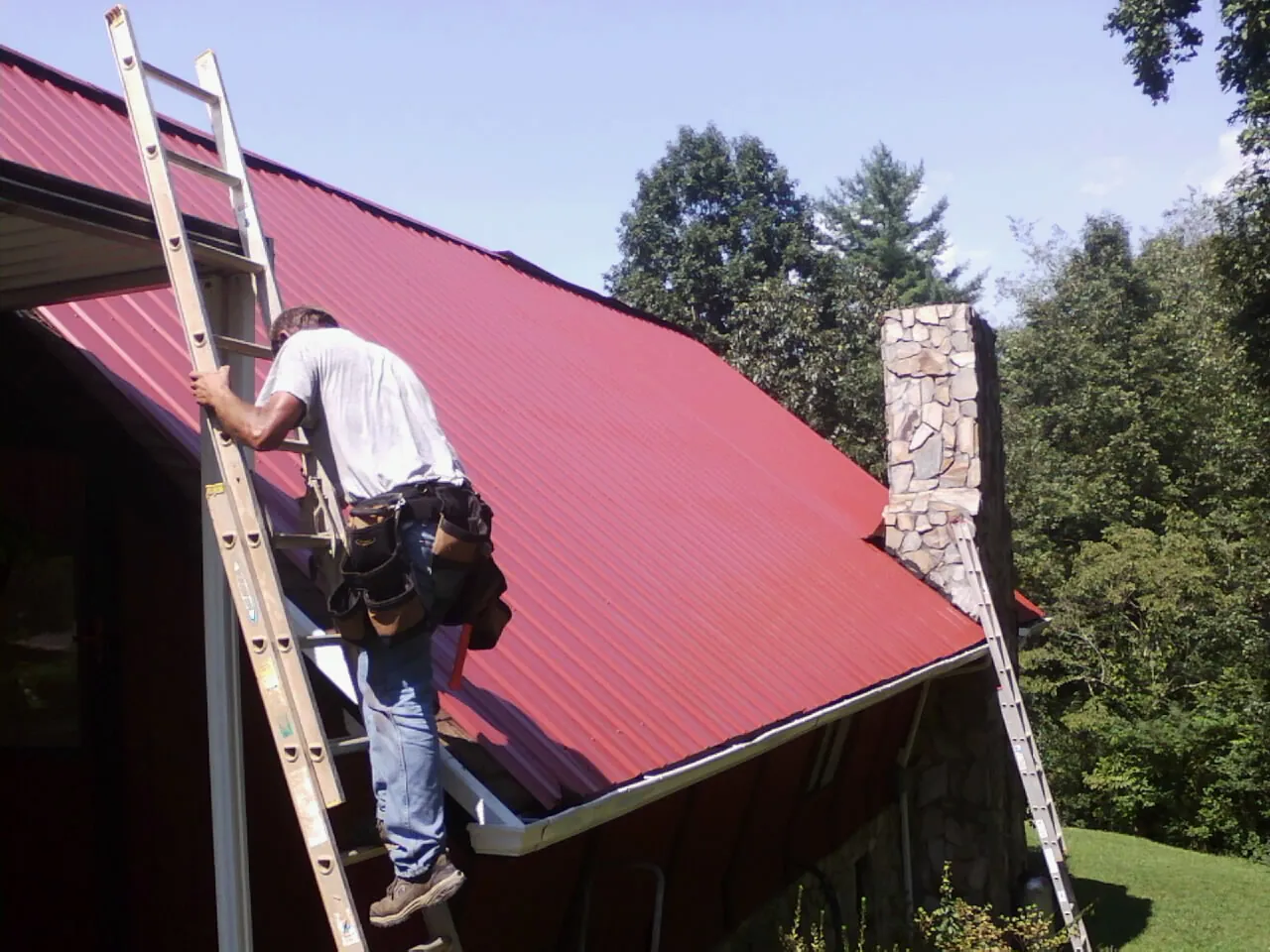 Roofer on a ladder beside a red metal roof and stone chimney — Soffit & Fascia Repair by Roofing Brothers in Robertsville